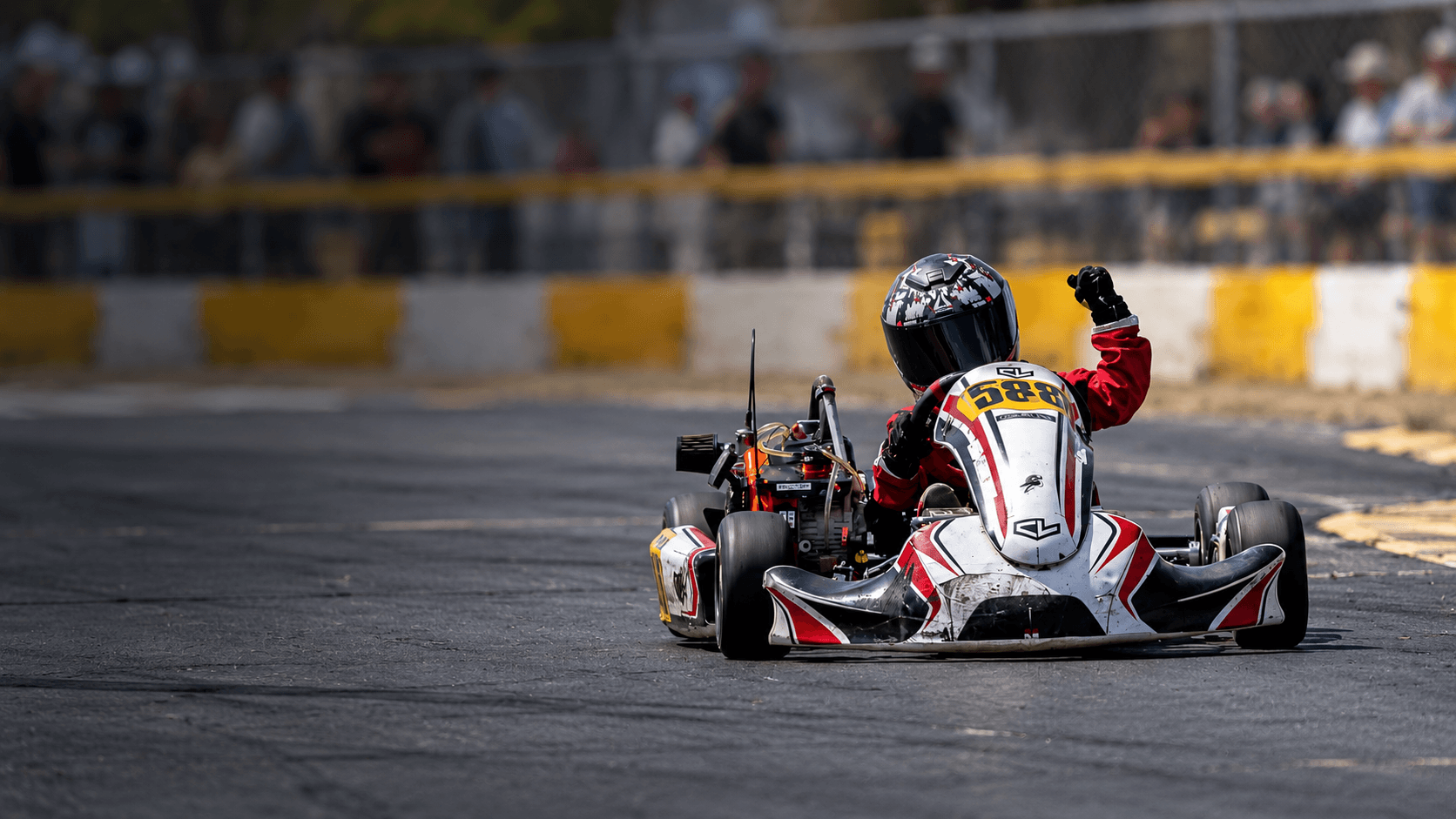 Young kart racer wearing a full-face helmet and red racing suit, raising a gloved hand while driving on an outdoor track.